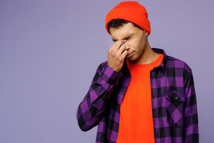 Young sad dissatisfed displeased man of African American ethnicity wearing casual shirt orange hat keep eyes closed rub put hand on nose isolated on plain pastel light purple color background studio.